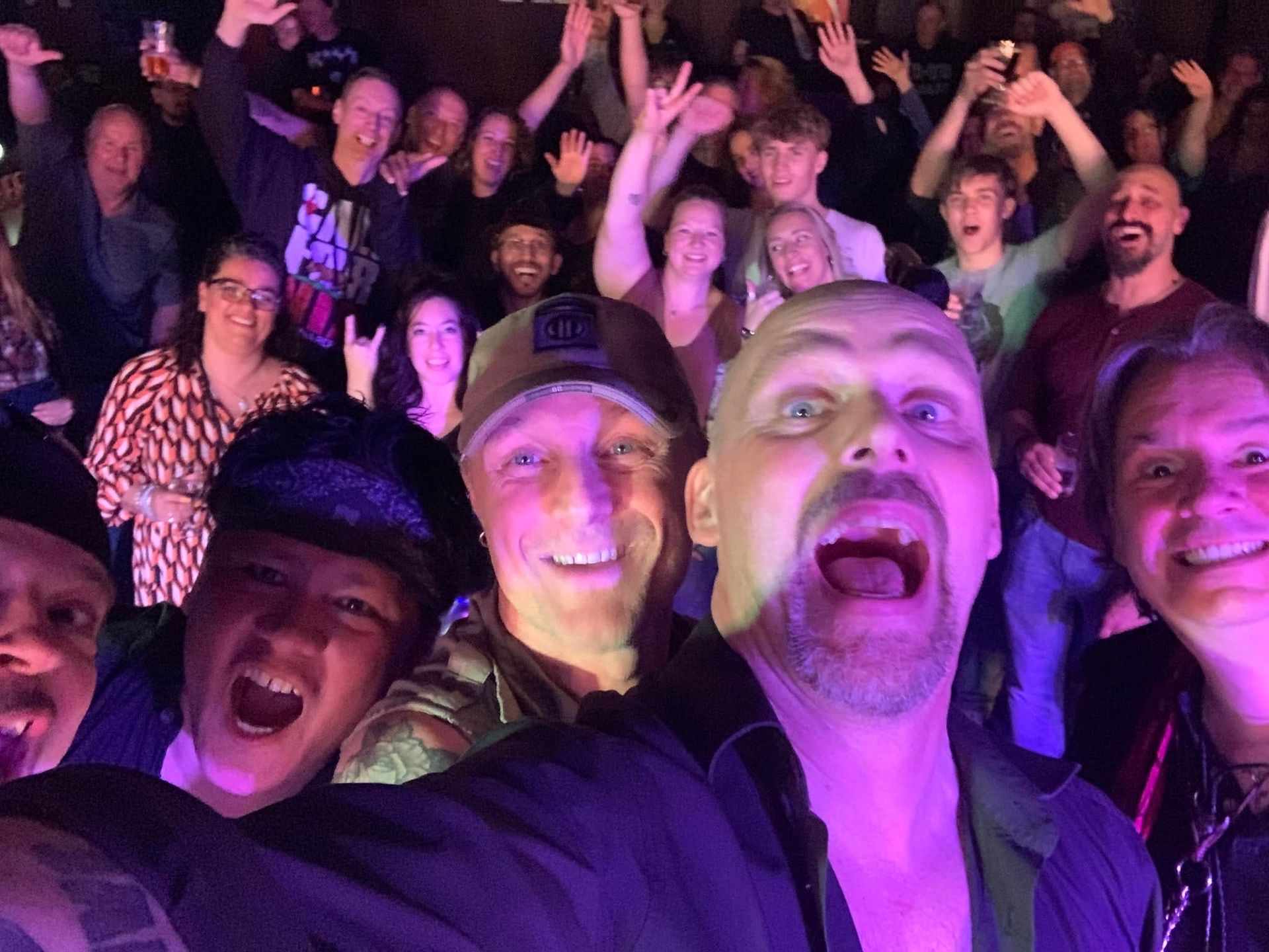 Publiek bij een concert juicht en lacht onder paarse podiumverlichting, met gezichten op de voorgrond die selfies maken.