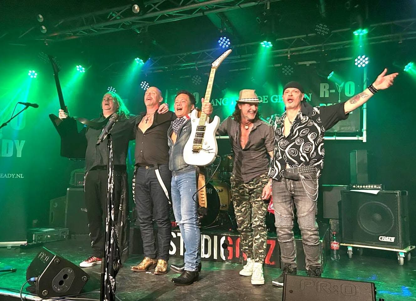Vijf artiesten staan ​​op een groen verlicht podium, lachend en zwaaiend, met een microfoon en gitaar in het zicht.