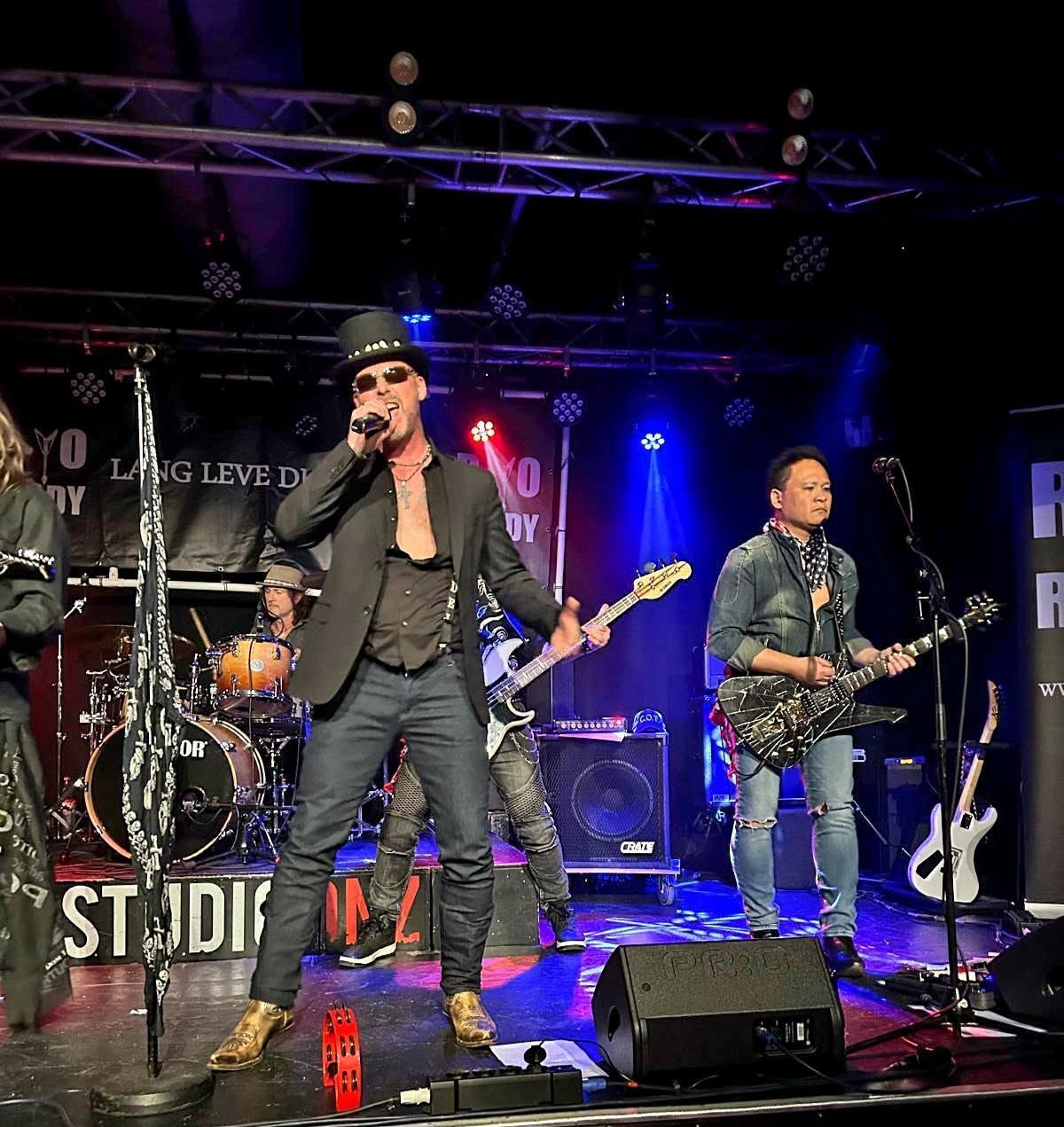 Rockband die optreedt op een verlicht podium, met een zanger met een zwarte hoed bij de microfoon in het midden en gitaren achter hem.