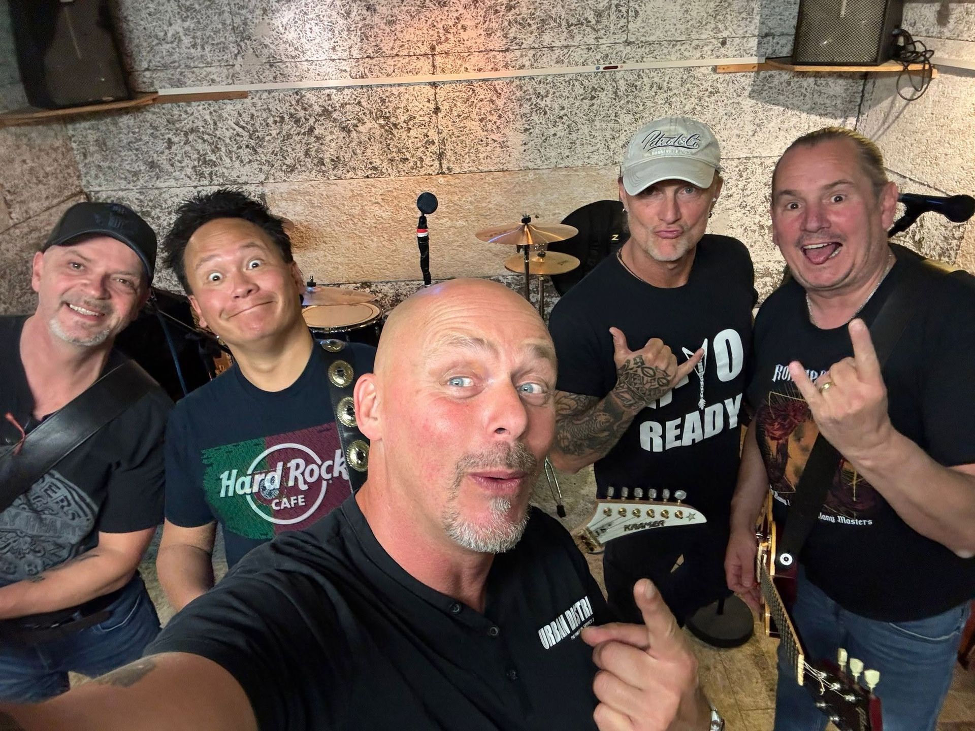 Vijf lachende bandleden poseren binnen met drums en instrumenten op de achtergrond.