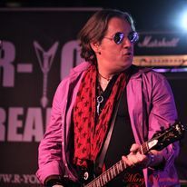 Muzikant in roze jasje en rode sjaal speelt gitaar op een verlicht podium.