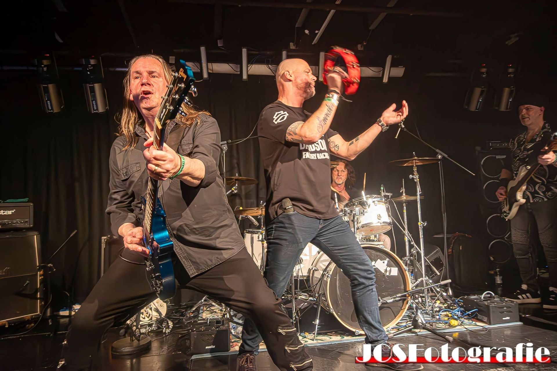 Rockband die optreedt op het podium, met gitaristen in donkere kleding onder podiumverlichting en een drummer op de achtergrond.