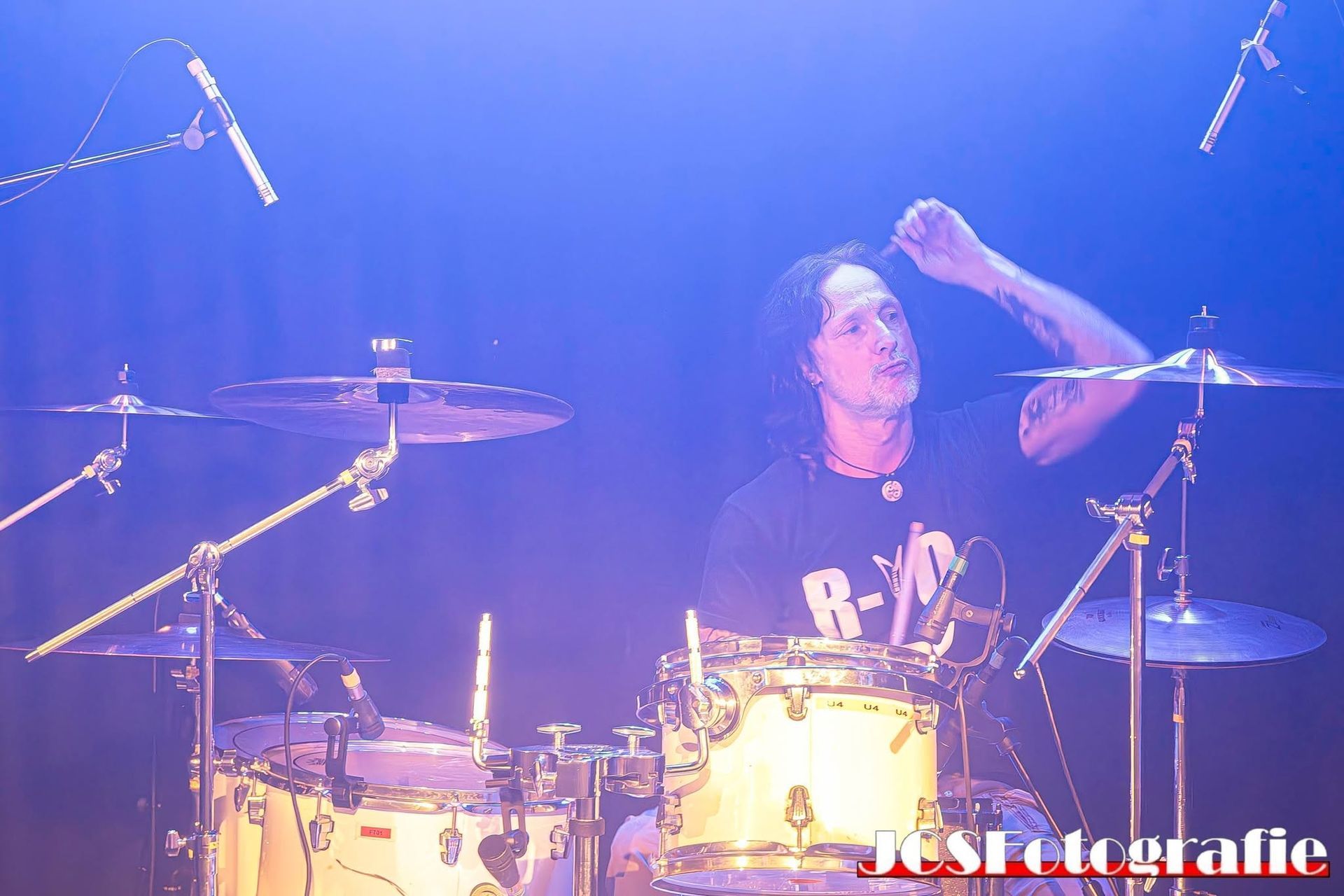 Een drummer treedt op het podium op onder blauwe en paarse lichten, terwijl hij een hand achter zijn drumstel omhoog houdt.