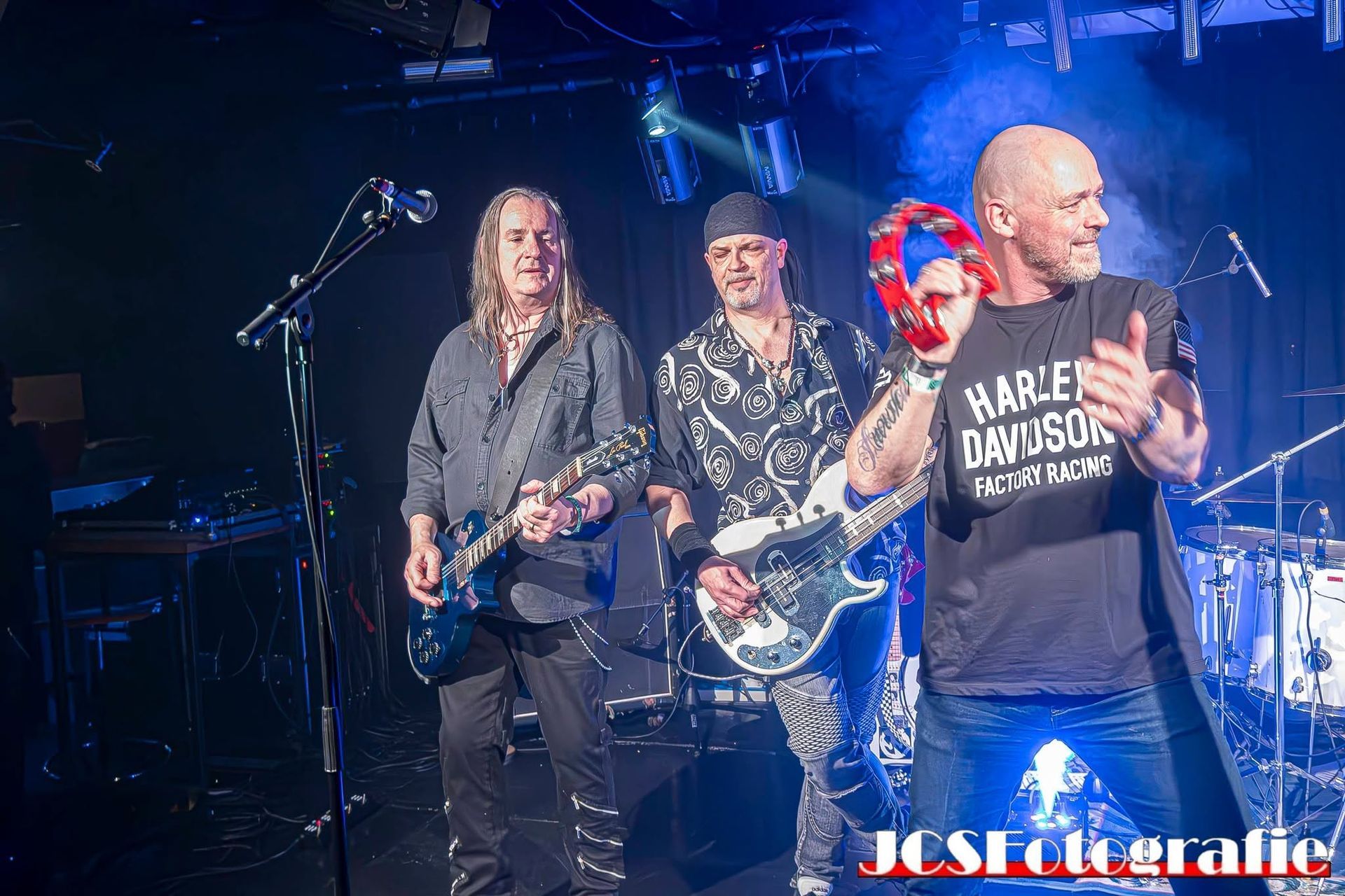 Rockband die optreedt op een blauw verlicht podium, met gitaren en een zanger in een Harley-Davidson shirt.