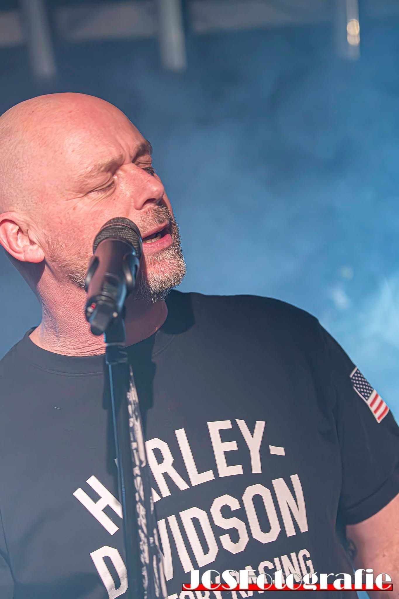 Zanger in een zwart Harley-Davidson shirt zingt in een microfoon, met blauwe podiumrook achter hem.