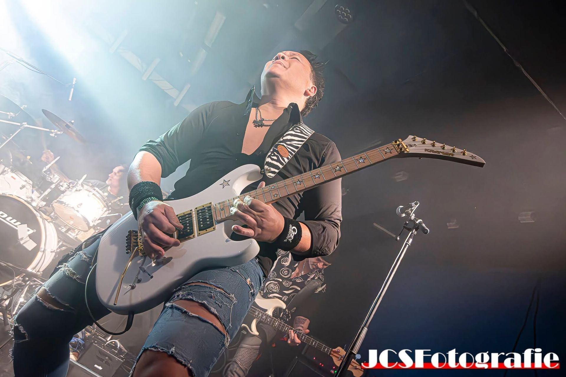 Gitarist speelt elektrische gitaar op een rokerig concertpodium onder felle lichten.
