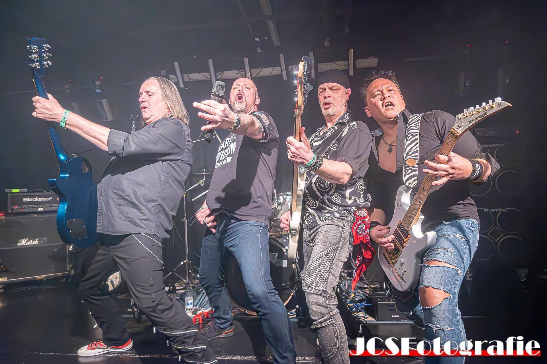 Rockband die optreedt op het podium met gitaren en dramatische belichting; een van de bandleden poseert links vooraan.