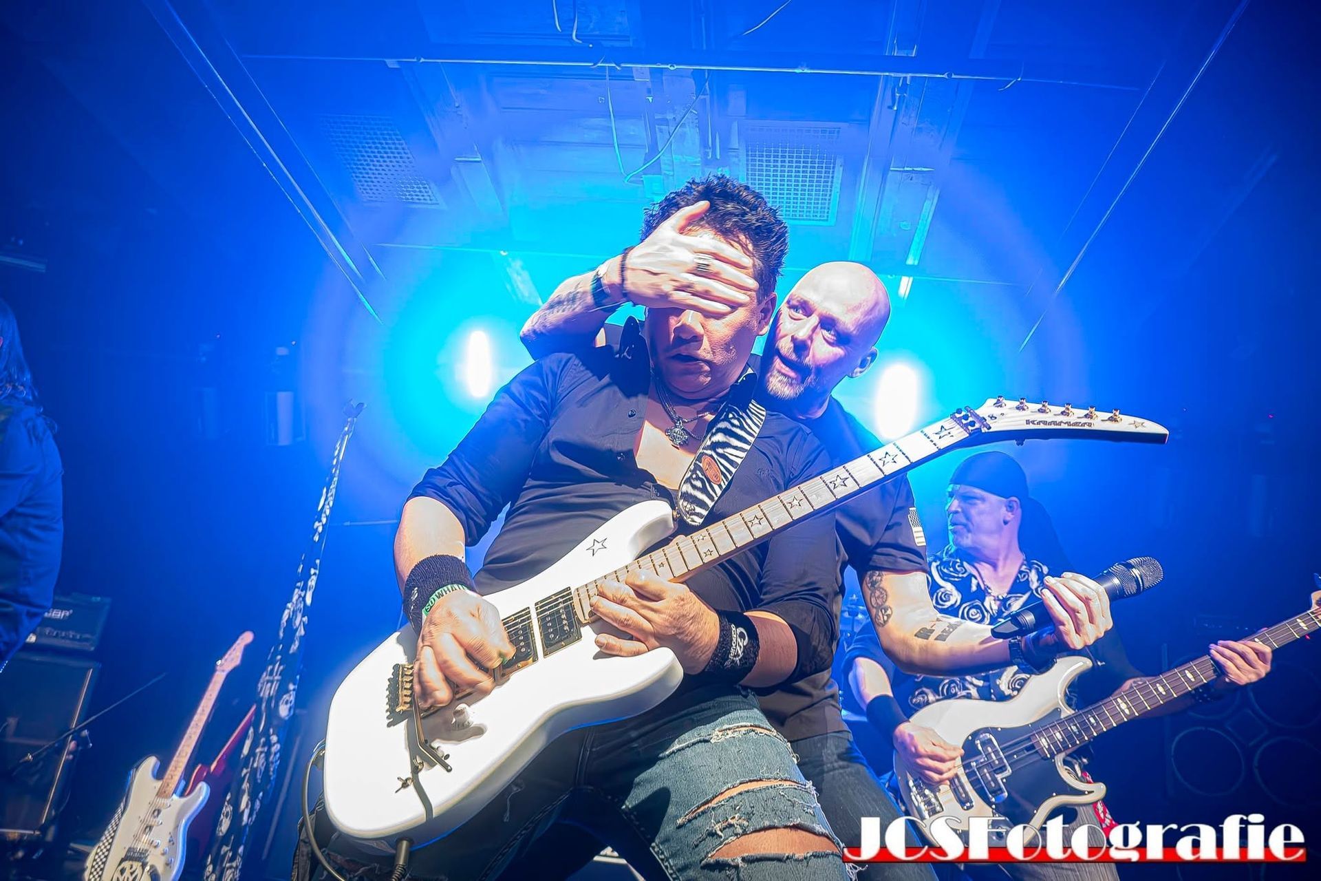 Rockband treedt op een podium op onder felblauw licht, gitarist op de voorgrond met een witte elektrische gitaar.