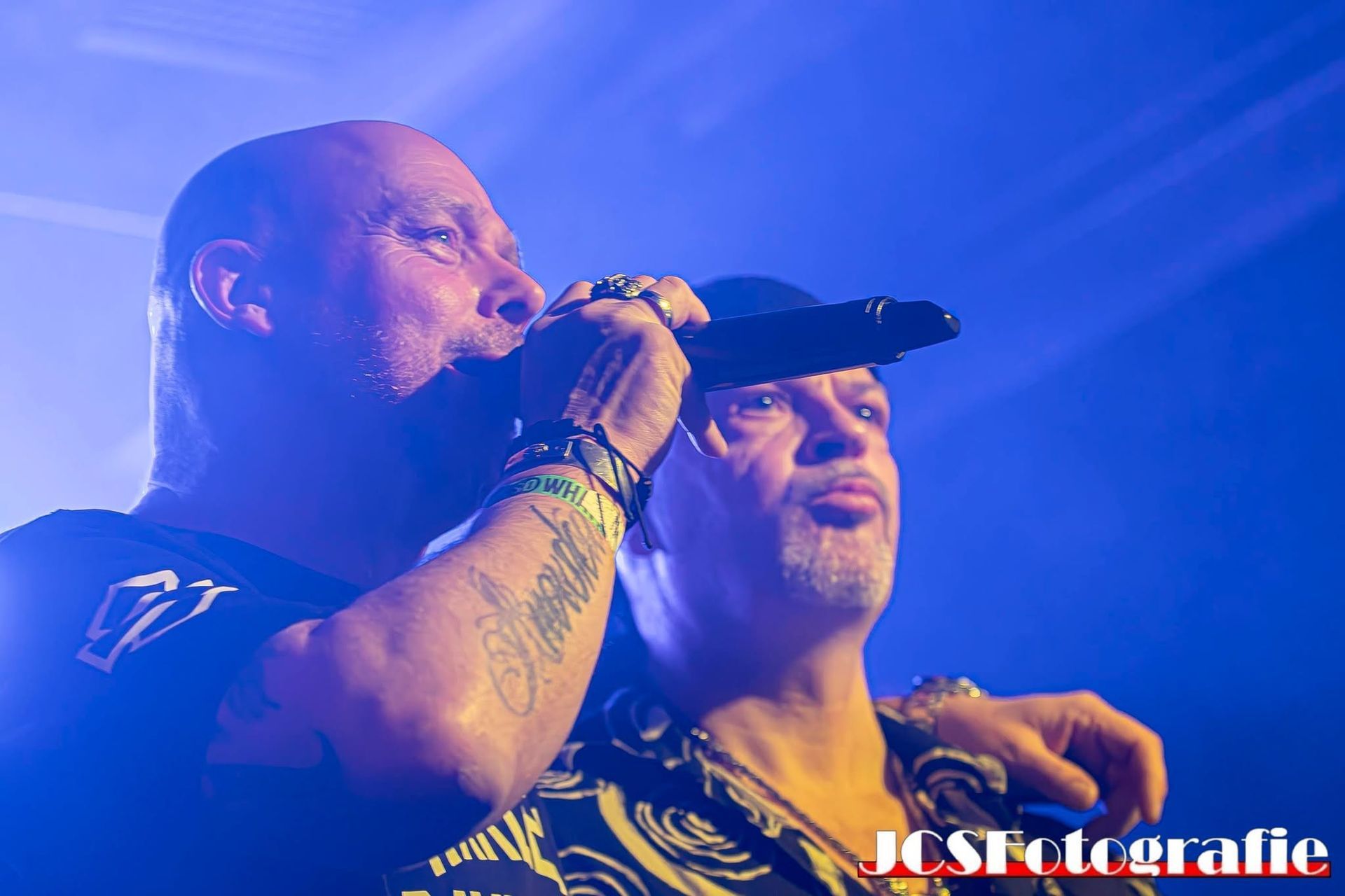Twee muzikanten staan ​​op het podium in blauw licht, de ene zingt in een microfoon terwijl hij arm in arm met de andere staat.