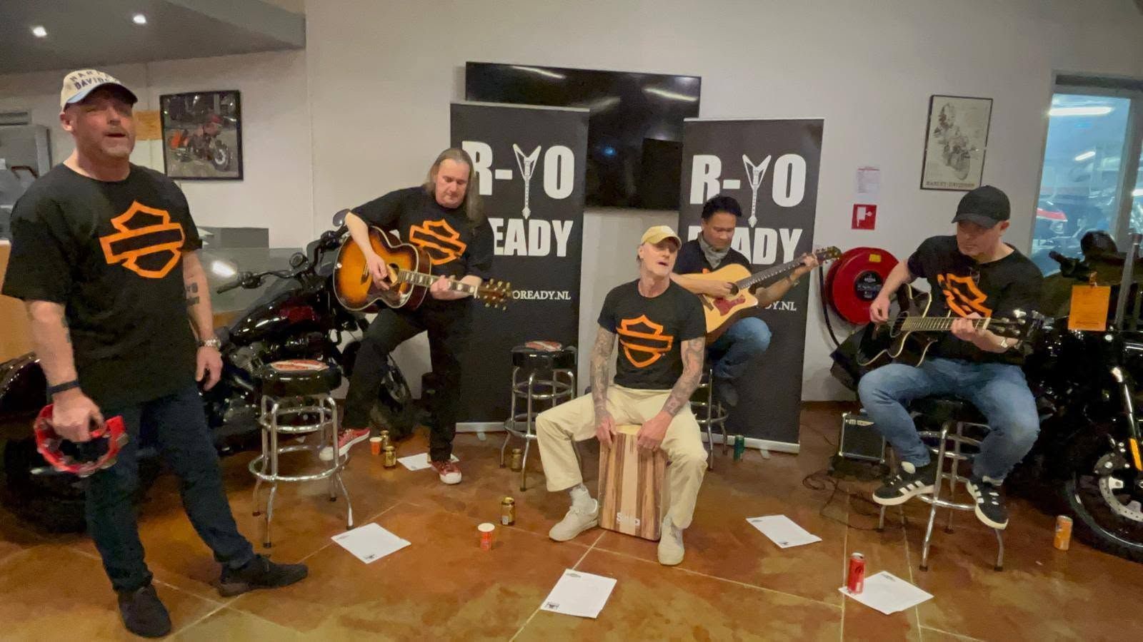 Vijf muzikanten spelen instrumenten in een woonkamer met zwart-oranje spandoeken op de achtergrond.