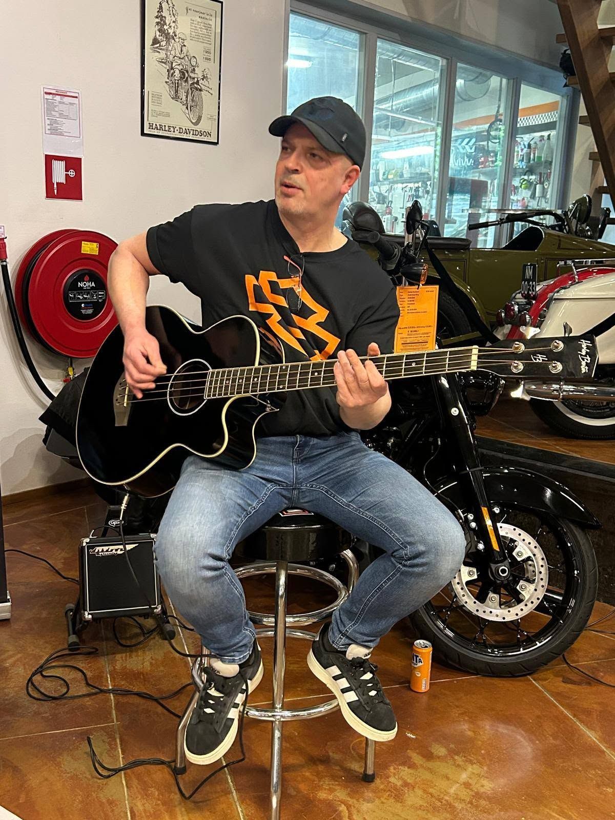 Een man zit op een kruk en speelt op een zwarte akoestische gitaar in een motorshowroom.