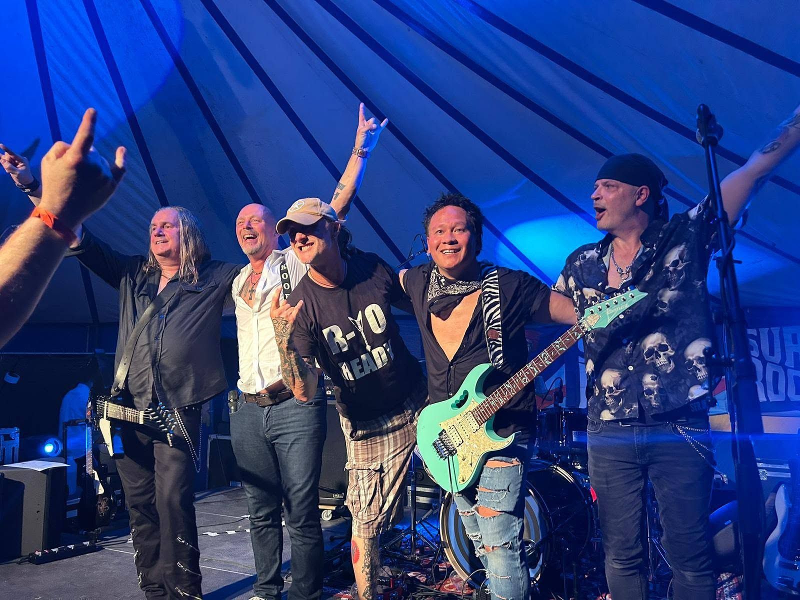 De bandleden poseren op het podium onder blauwe lichten, lachend met hun gitaren omhoog tijdens een live optreden.