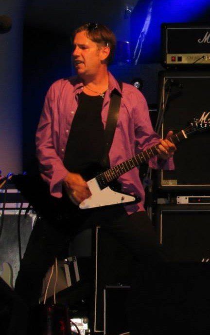 Muzikant in een roze shirt die elektrische gitaar speelt op een schemerig verlicht podium.