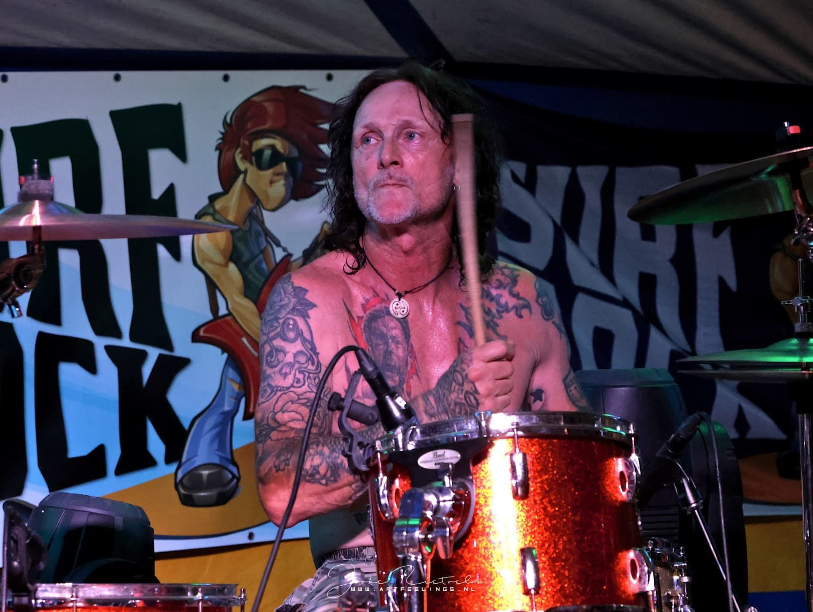 Drummer die optreedt op het podium achter een oranje drumstel met een rockbanner op de achtergrond.