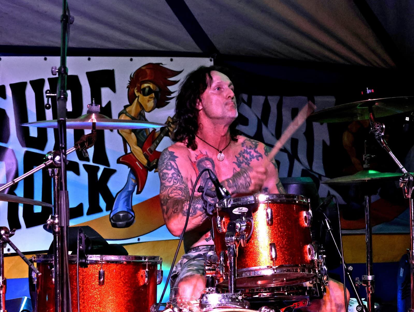 Een drumster speelt op het podium onder paarse verlichting met een oranje drumstel en een rockbanner achter haar.