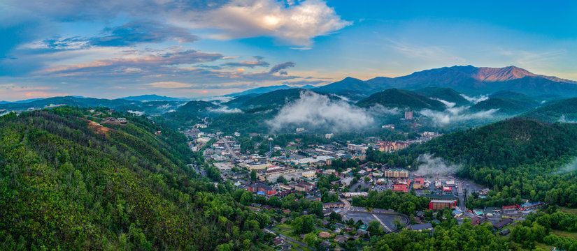 Riverfront town at sunset with a bridge, grassy bank, and buildings under a cloudy sky in Gatlinburg, TN.