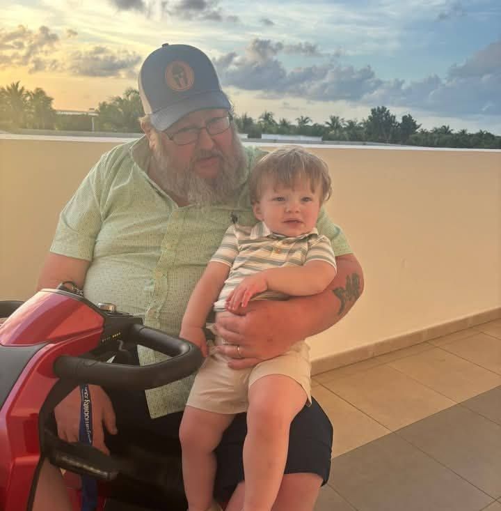 Client Mikey Justus is holding a grandchild on his lap while riding a red scooter at sunset on a tiled balcony. 