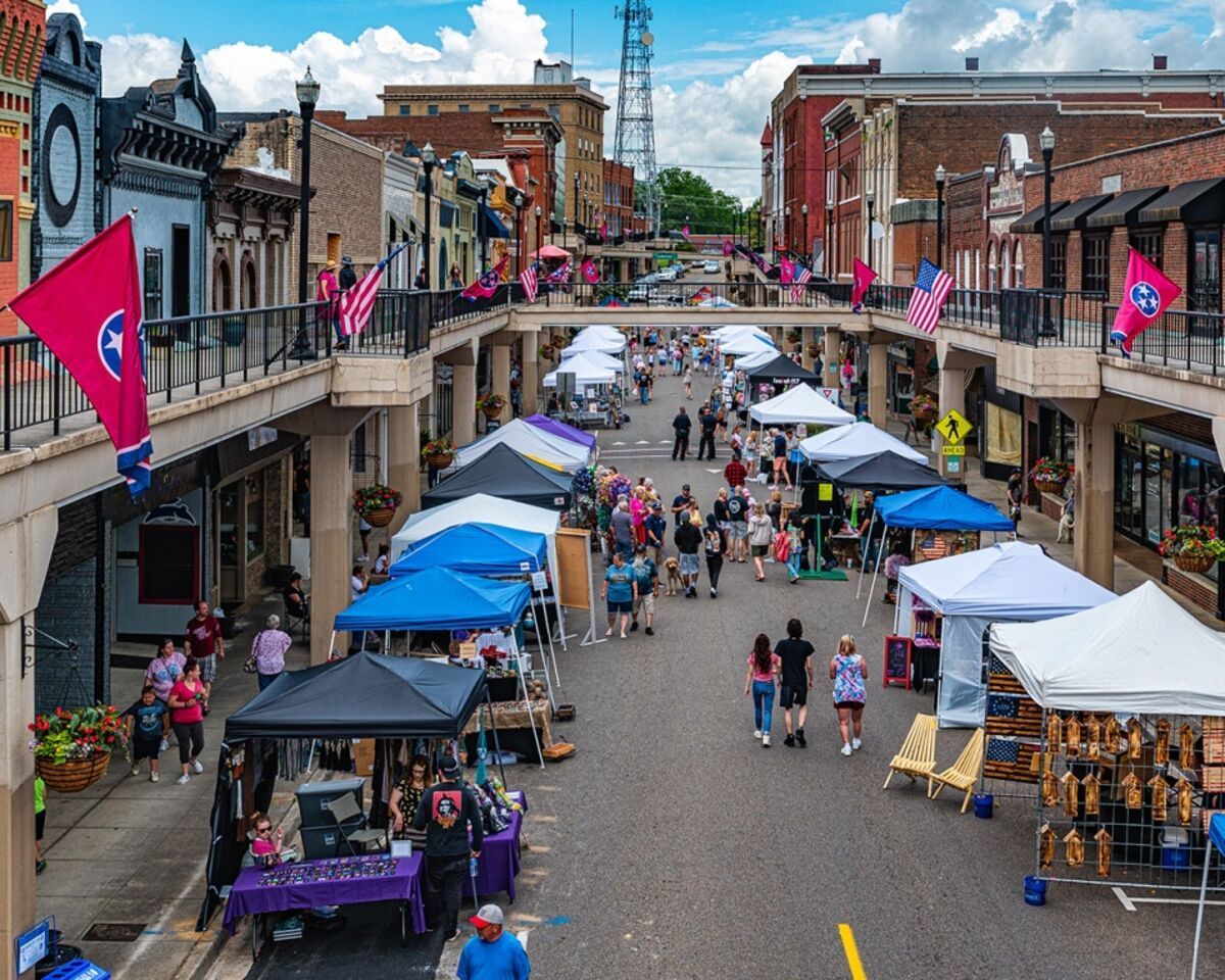 Outdoor market on a Morristown, TN city street with tents, pedestrians, brick buildings, and pink flags under a cloudy sky.
