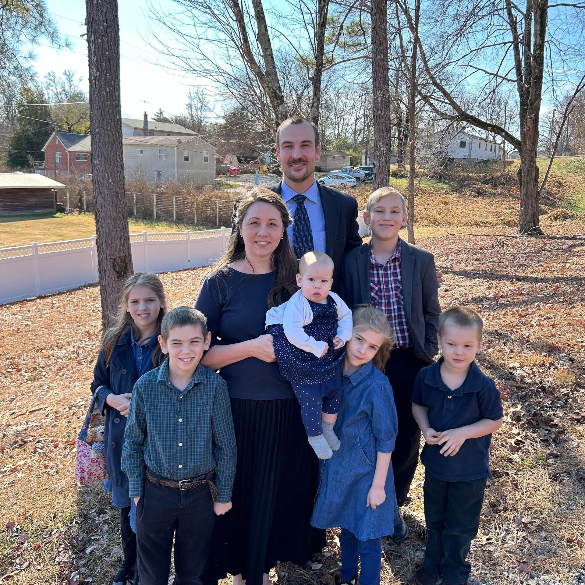Family of seven posing outdoors in a sunny yard, with a baby in front and trees and houses behind with client Stephen Easley in the back behind his family. 