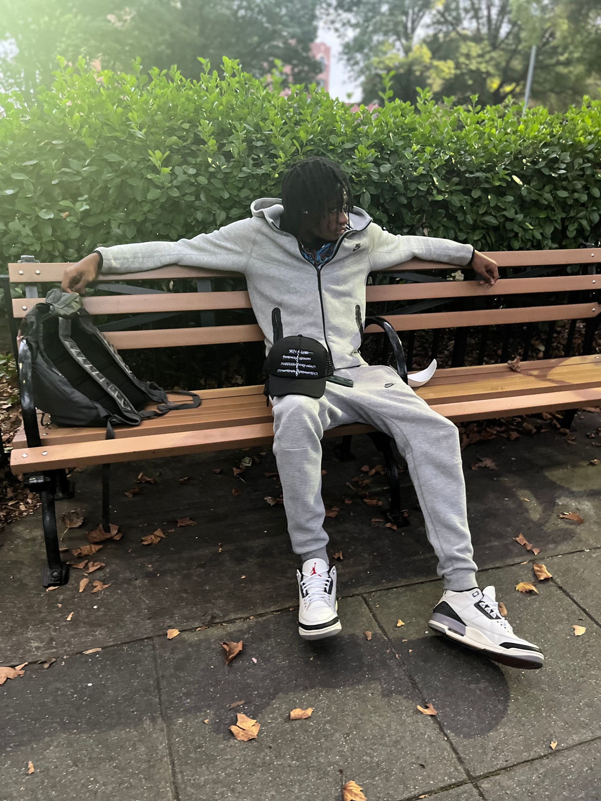 Person in gray sweatsuit sits on park bench with backpack, small bag, and headphones.