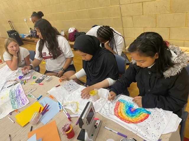 Group of people at a table, drawing on fabric with markers and other art supplies in a room.