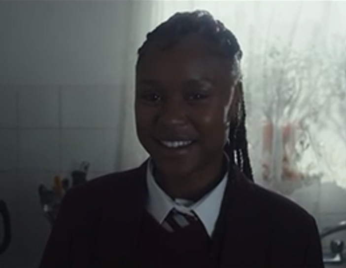 Girl in school uniform smiling, standing in a kitchen with window and sink in the background.