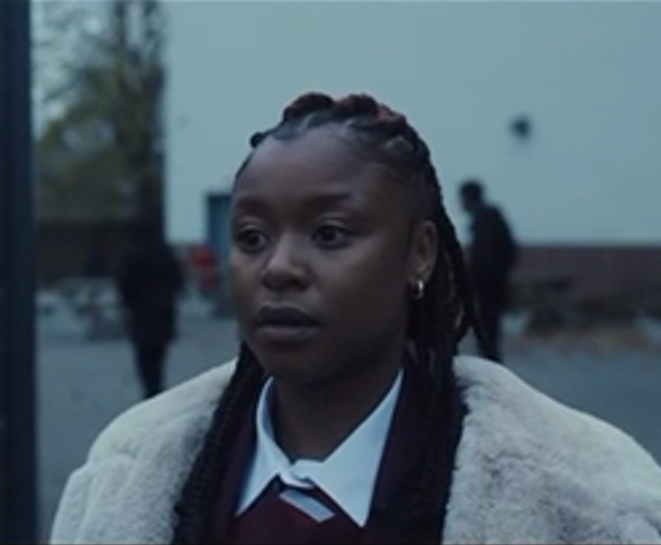 A person with braided hair wearing a white fur coat and collared shirt looks concerned outdoors.