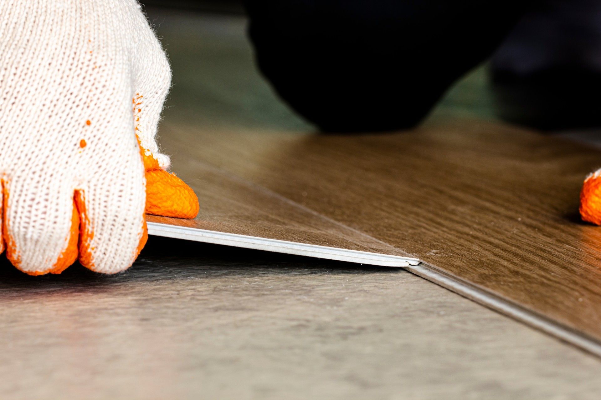 A person wearing orange gloves is cutting a piece of vinyl flooring.