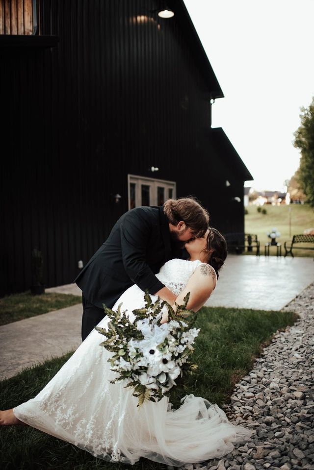 Bride and groom kissing outdoors beside a dark barn, with the bride in a white gown and gravel path nearby