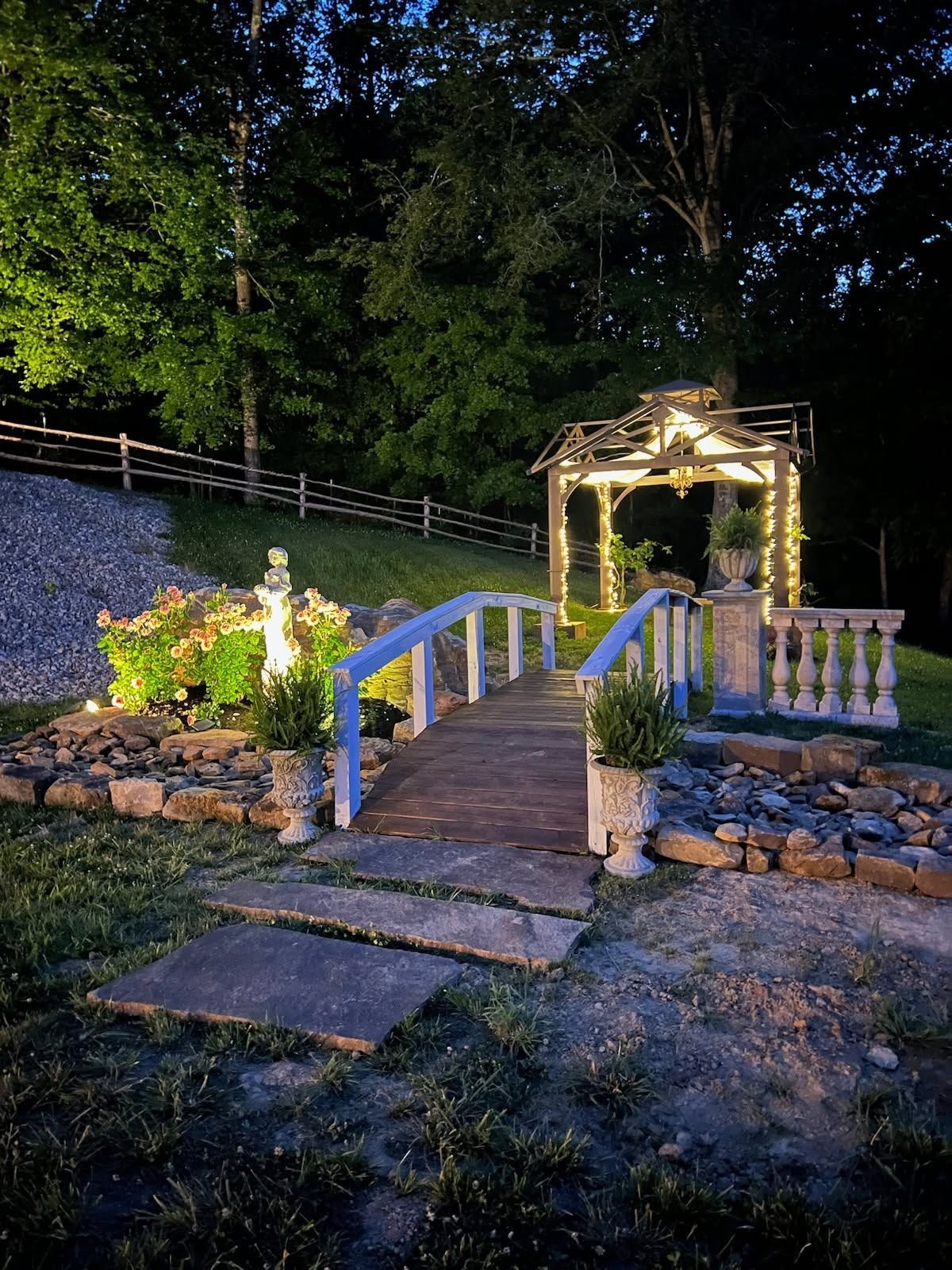 Lit gazebo and wooden bridge in a landscaped garden at dusk, with stone walls and trees.