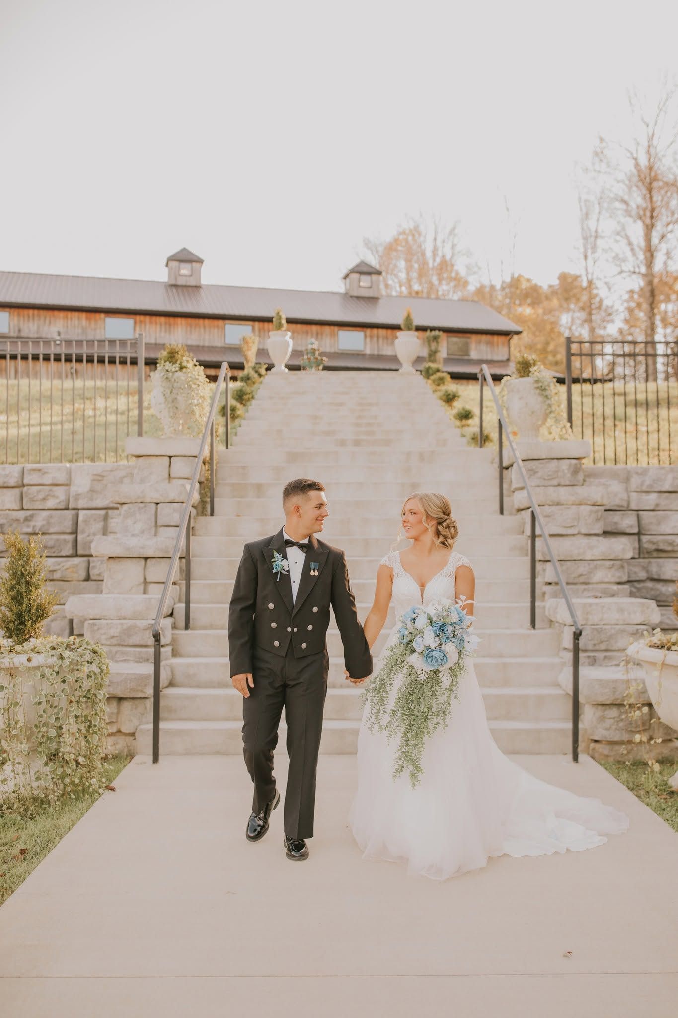 Bride and groom walking down a stone path at an outdoor wedding venue, holding a blue-and-white bouquet