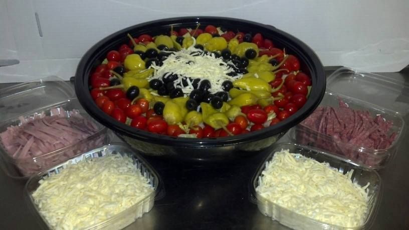 Prepared salad with tomatoes, peppers, olives, and cheese, surrounded by containers of meat and cheese.