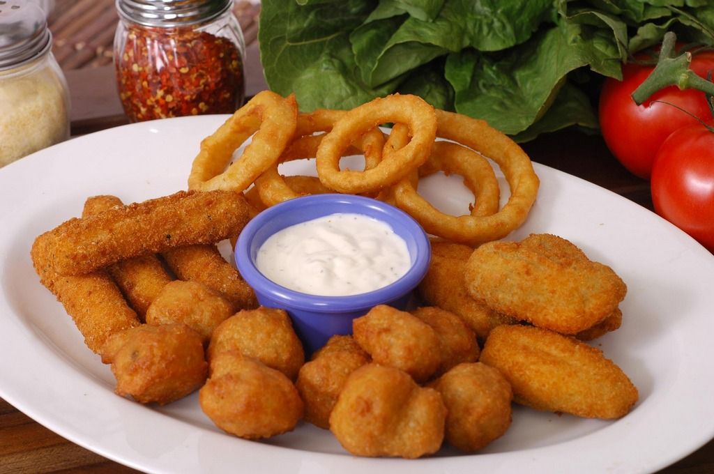 Plate of fried food: onion rings, mozzarella sticks, mushroom bites, dipping sauce. Tomatoes and greenery visible.