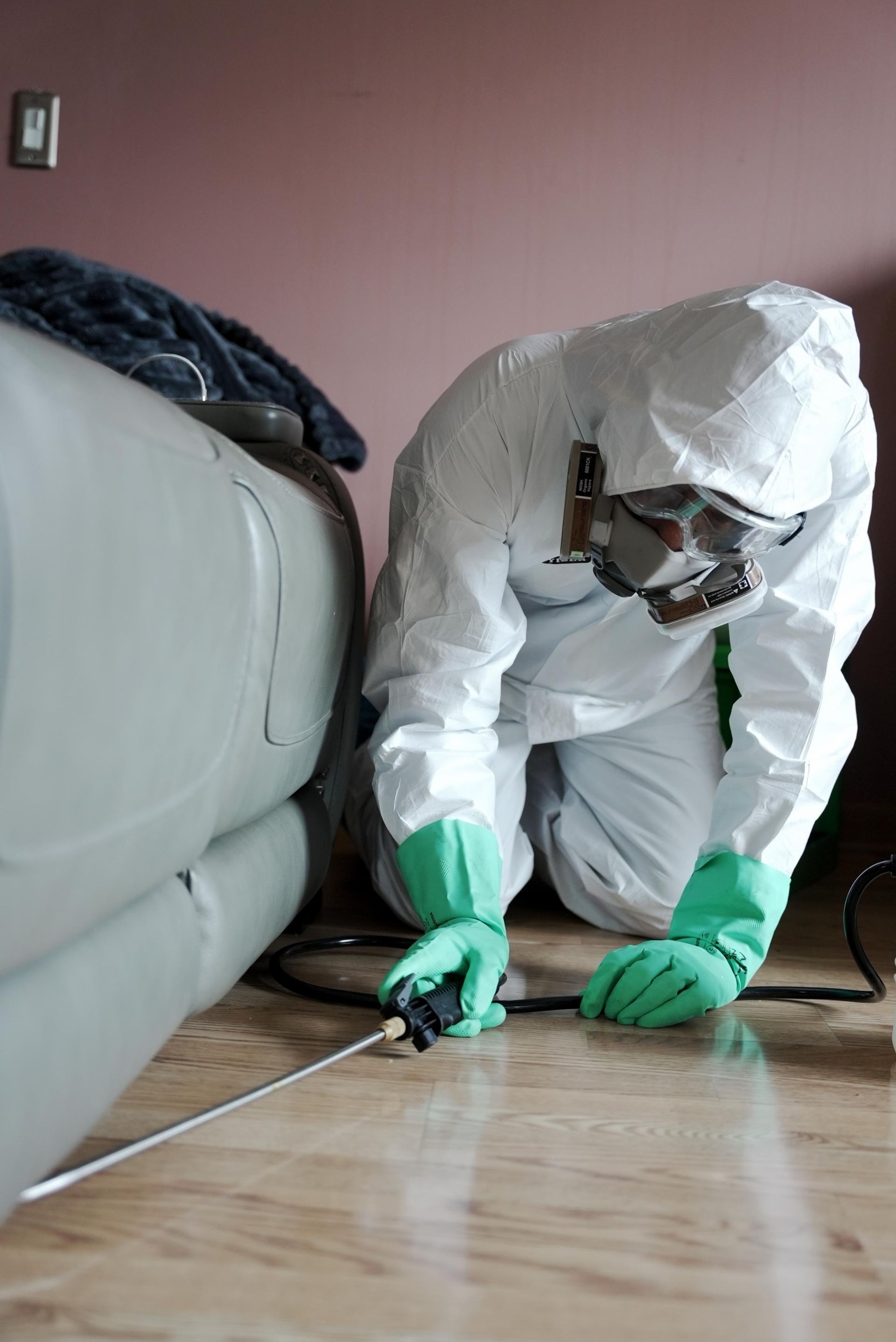 Person in a white protective suit and green gloves cleaning a floor beside a couch.