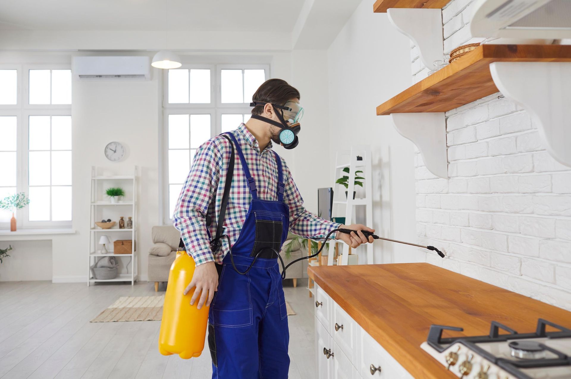 Worker in blue overalls disinfects a kitchen countertop while holding a yellow spray bottle.