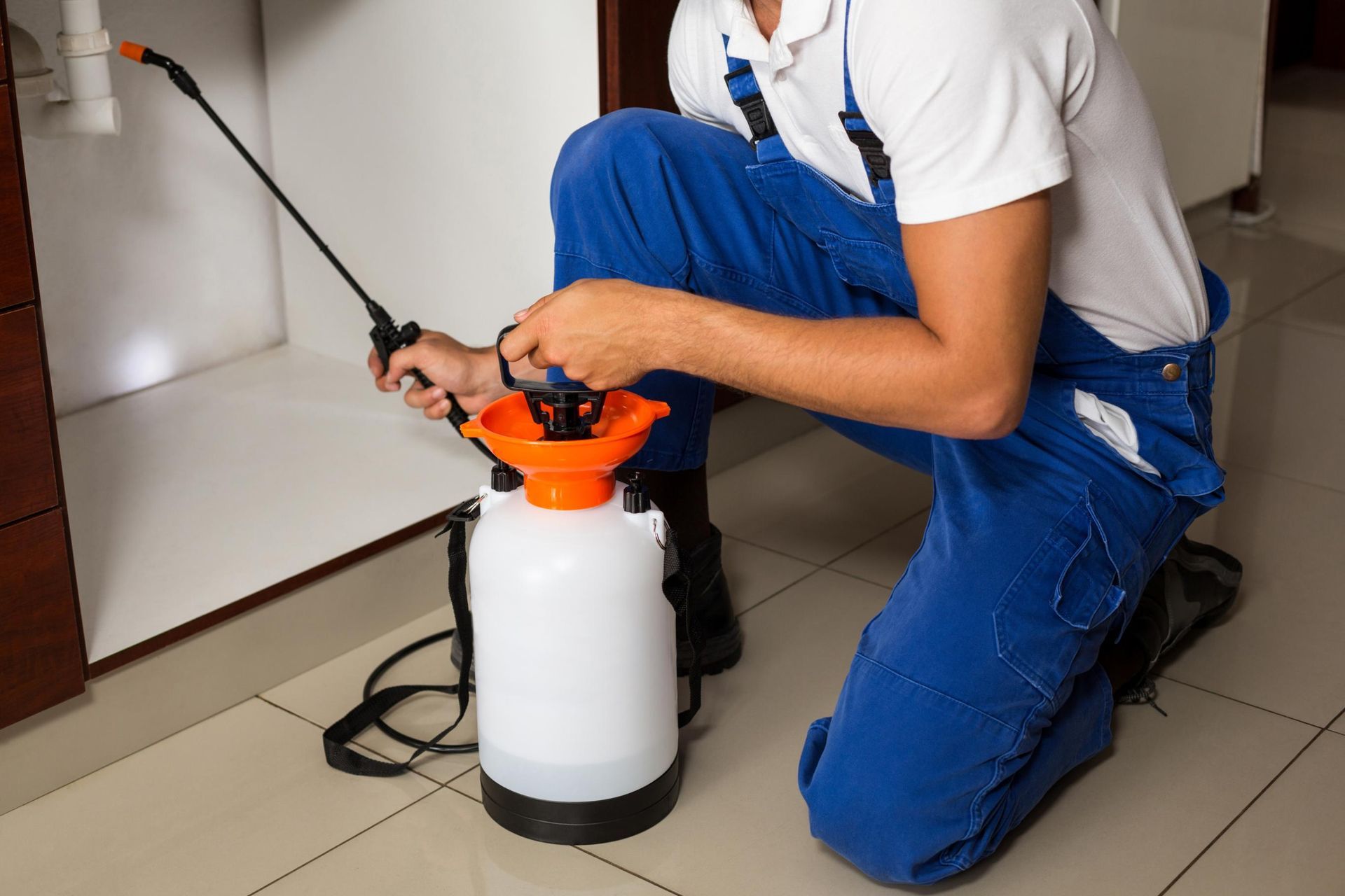 Person using a backpack sprayer indoors on a tiled floor, wearing blue overalls and kneeling.