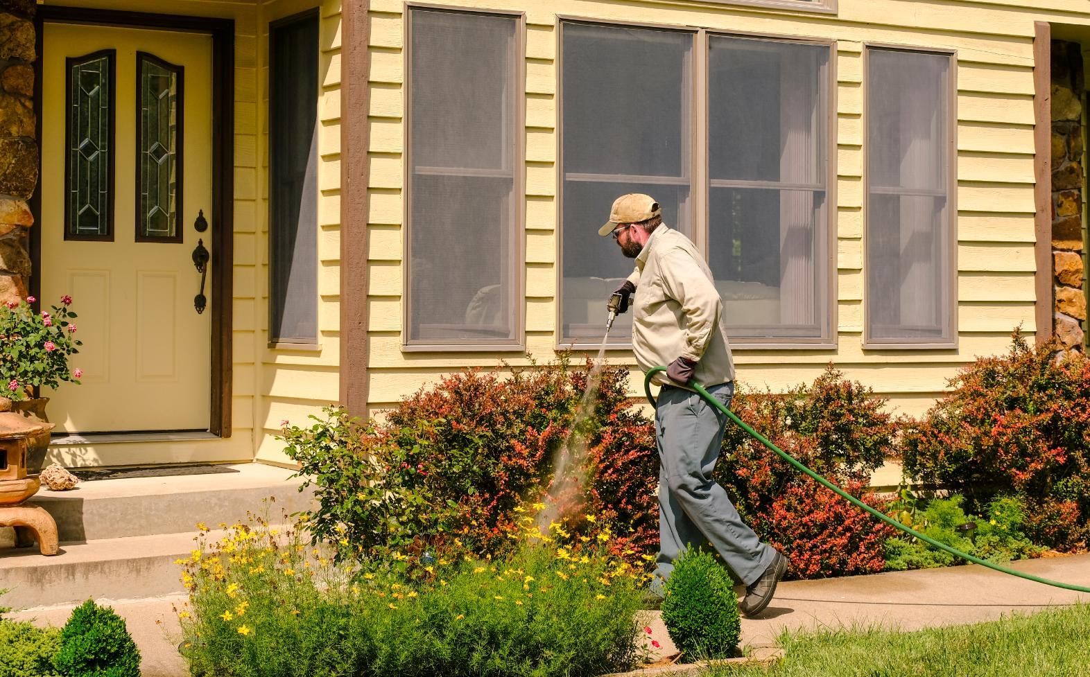 Person pruning bushes in front of a beige house with blue shutters and a flower bed