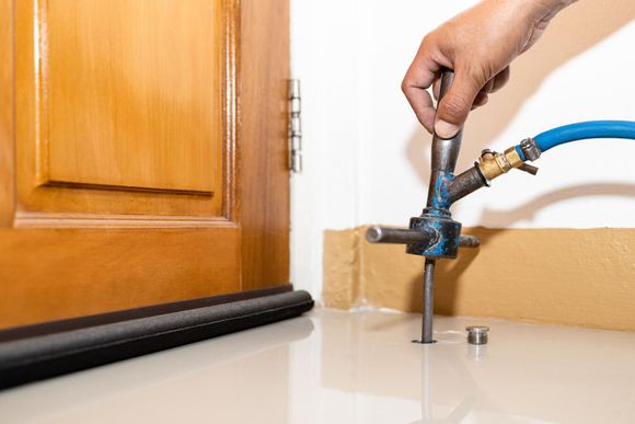 Hand using a hose to spray water at the base of a wooden door and wall on a floor.