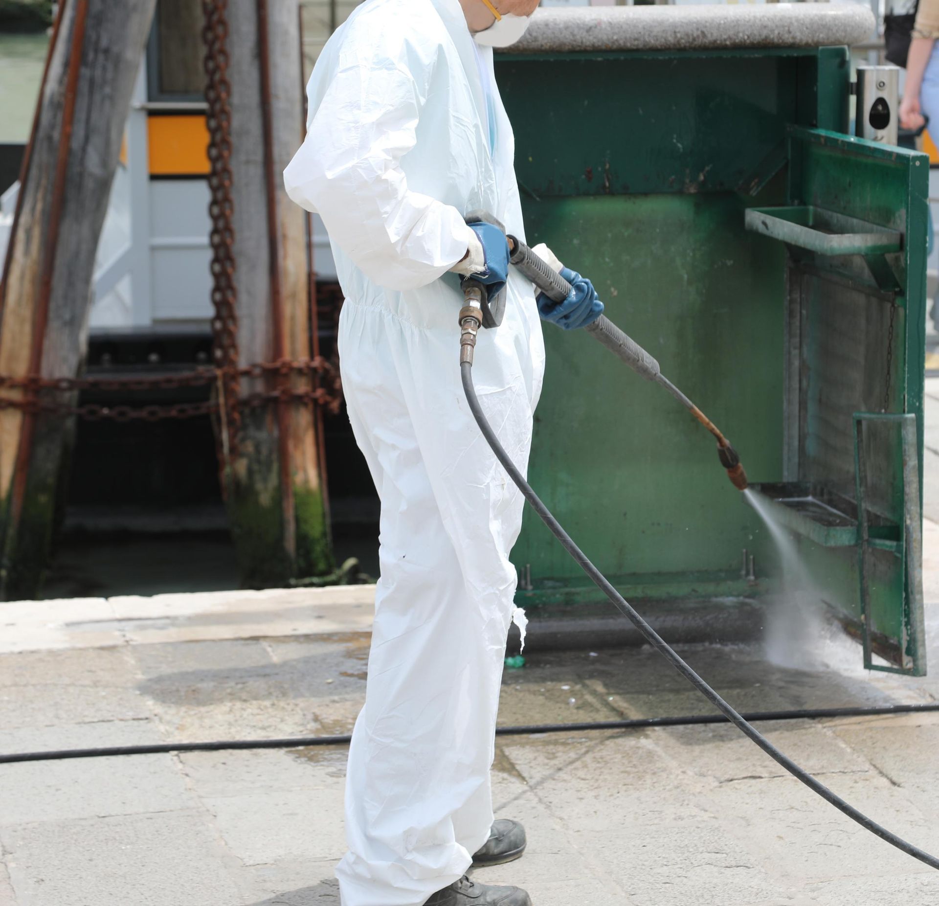 Worker in white protective suit pressure-washing a green industrial surface outdoors