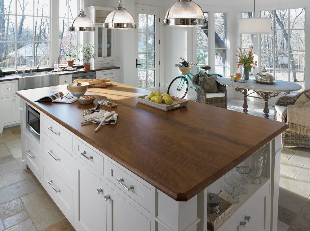 Bright modern kitchen island with wood countertop, white cabinets, pendant lights, and bicycle by large windows