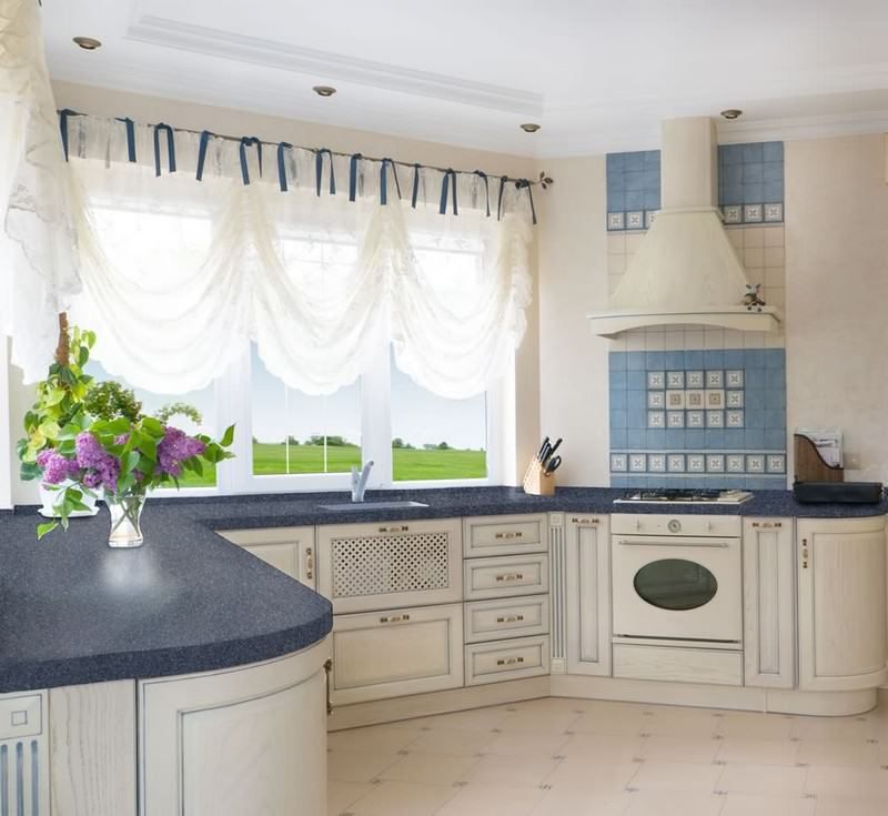 Bright white kitchen with blue accents, curtained window, dark countertop, and vase of flowers