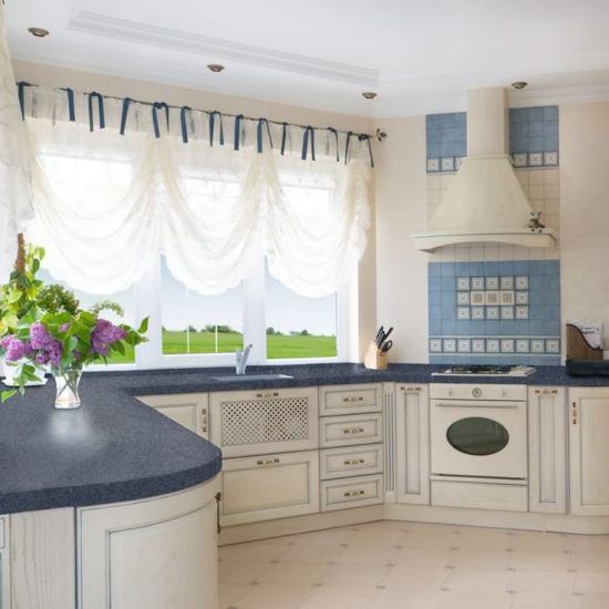 Bright cream kitchen with blue countertops, tiled backsplash, and a large window with white curtains and flowers.