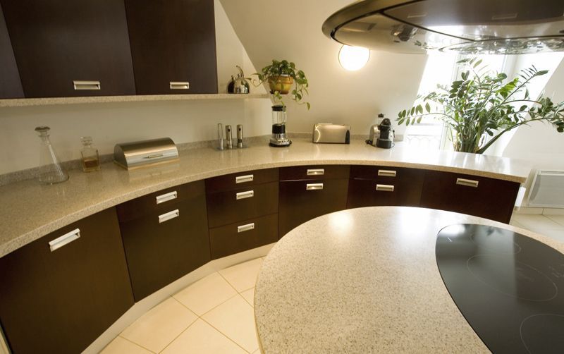 Modern kitchen with dark cabinets, curved granite countertop, tiled floor, and bright windows with plants