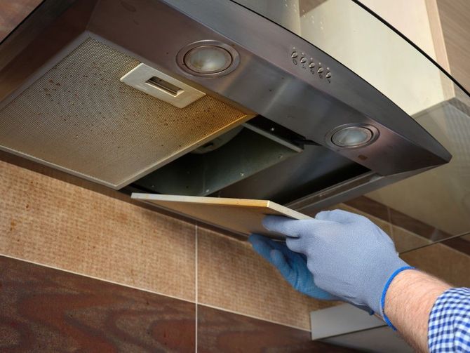 Gloved hand removing a vent cover from a kitchen range hood over a tiled backsplash