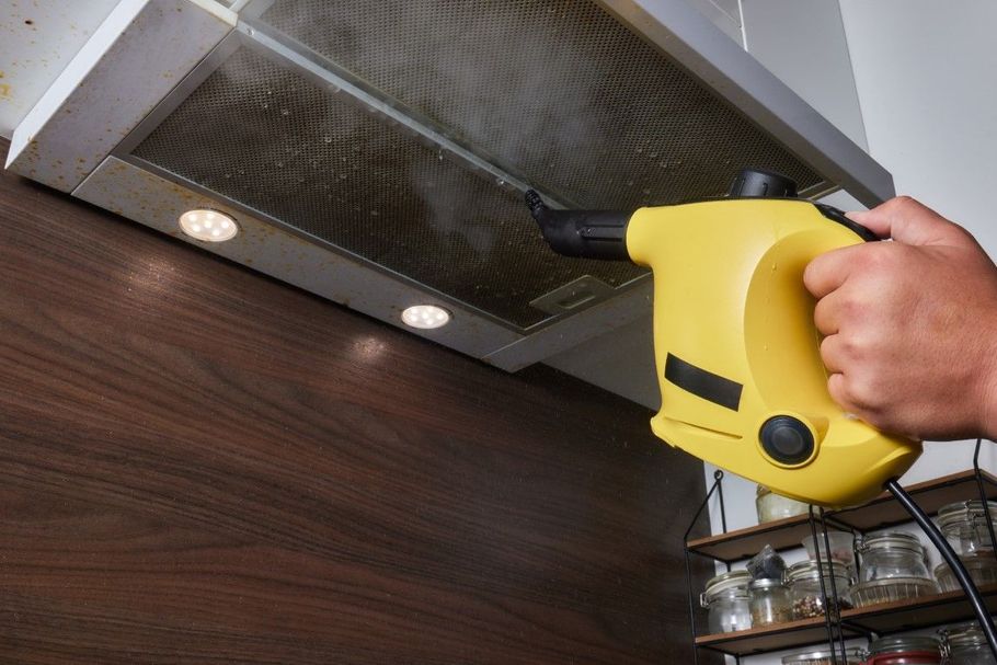Handheld yellow steam cleaner cleaning a metal kitchen range hood under cabinet lights