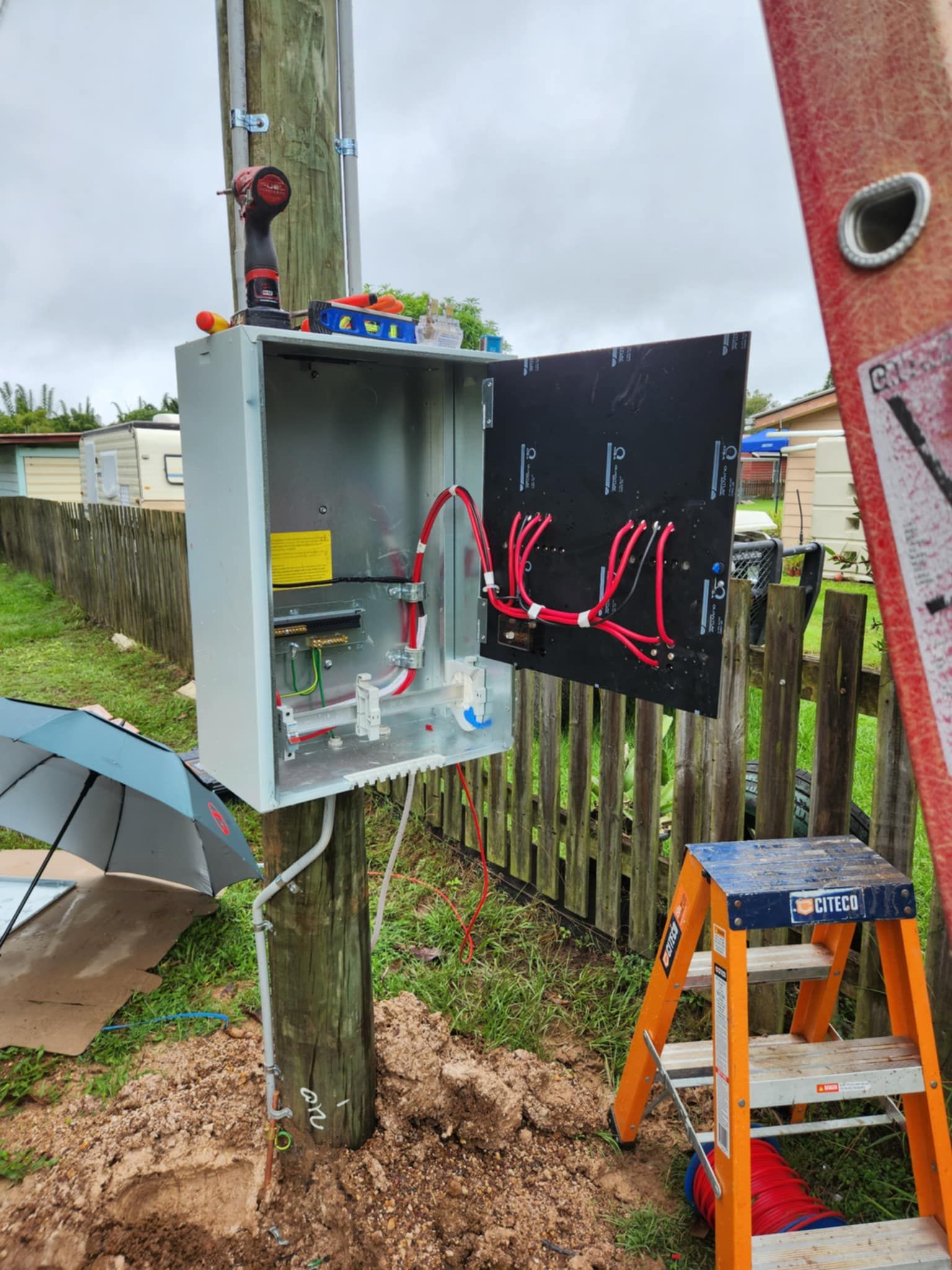 Electrical box mounted on a pole; open door reveals wiring. A ladder, drill, and umbrella are nearby — Watkins Electrical In Pialba, QLD