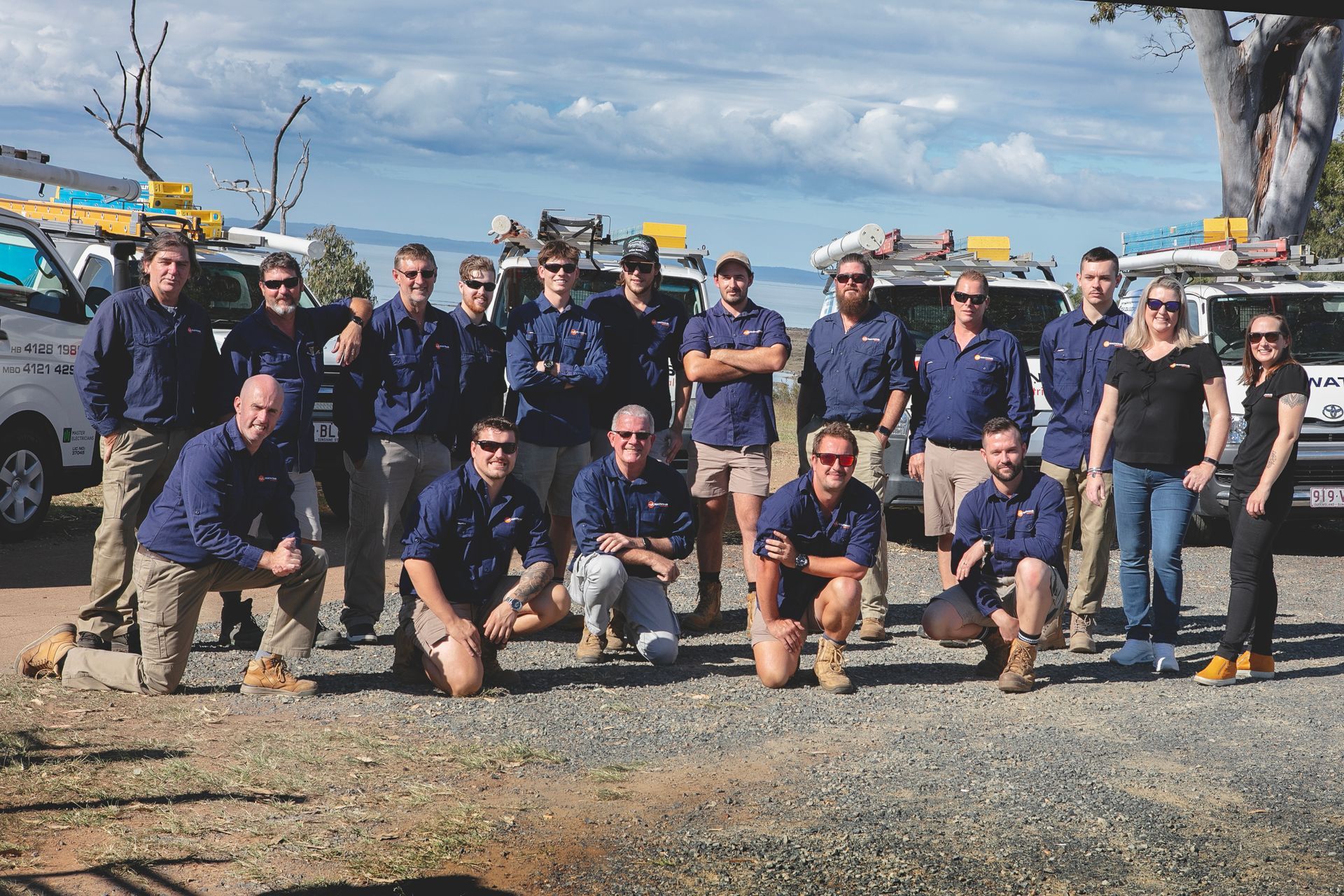 Group of workers in blue shirts and khaki shorts posing with company vans outdoors — Watkins Electrical In Pialba, QLD