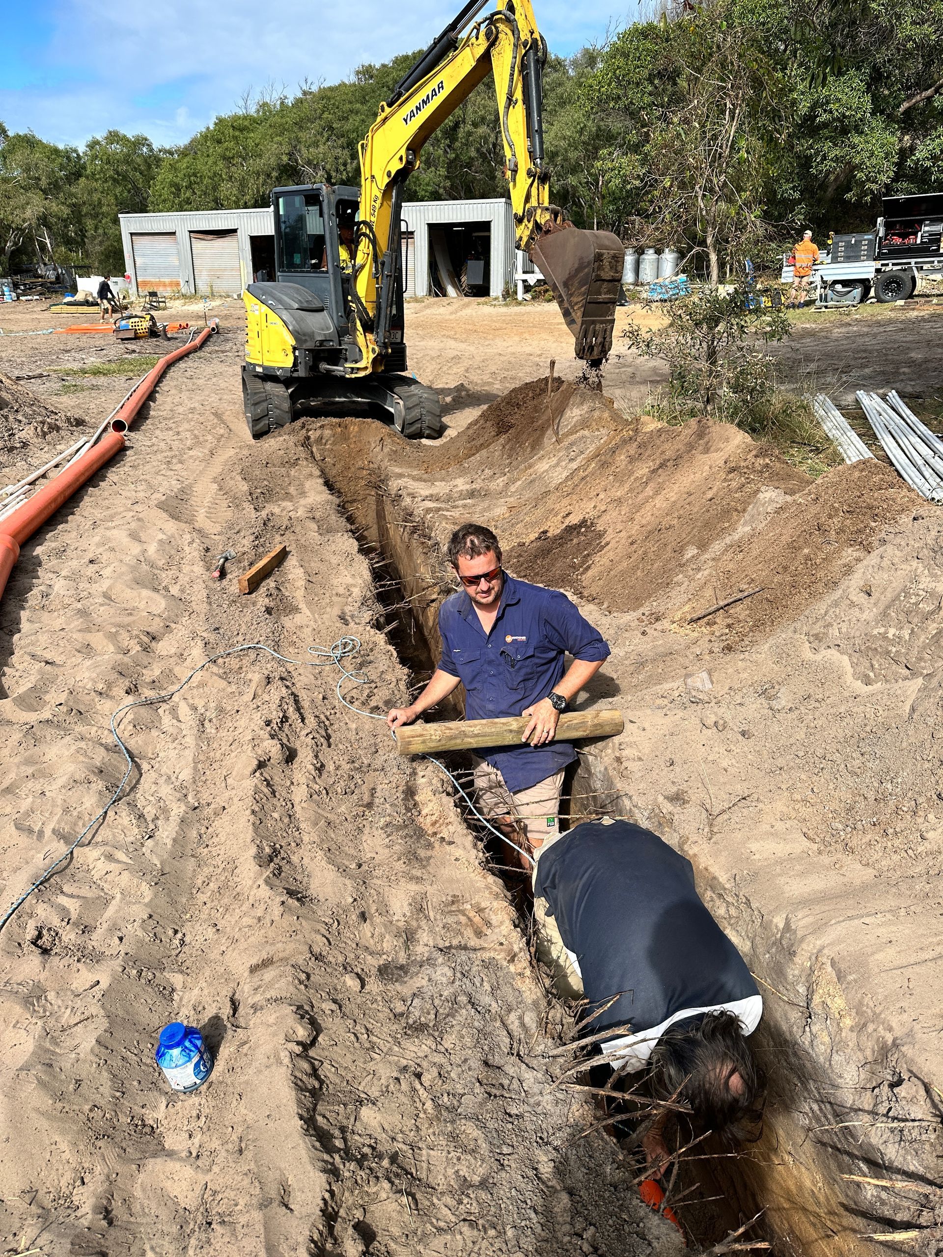Workers installing a pipe in a trench with an excavator in the background at a construction site — Watkins Electrical In Pialba, QLD