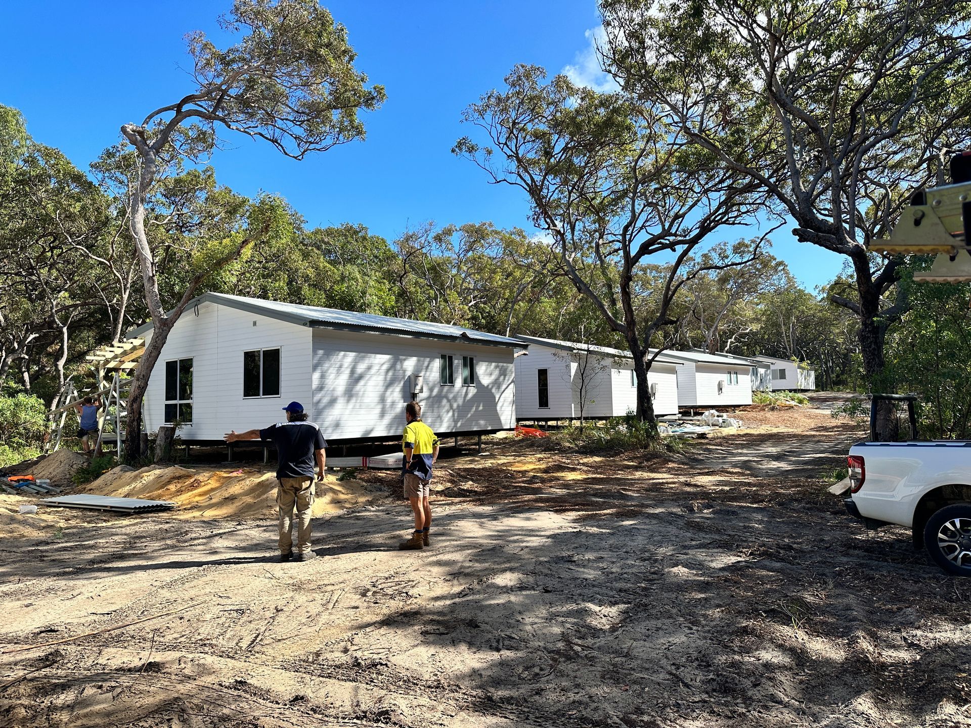 People walking toward a row of white buildings on a dirt road, trees on either side, under a blue sky — Watkins Electrical In Pialba, QLD