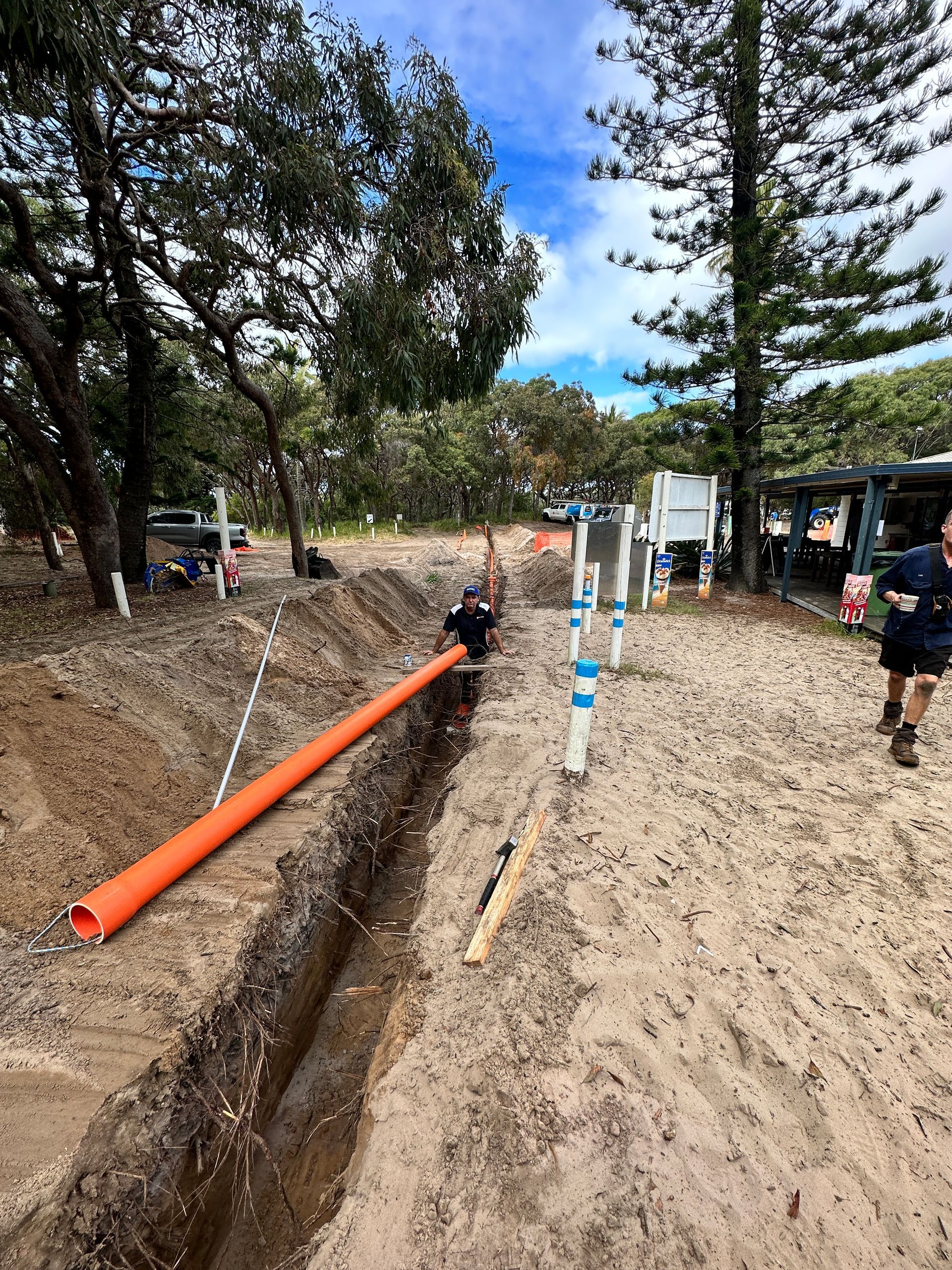 Trench with orange pipe in sandy soil, blue pipes, person walking, beach setting — Watkins Electrical In Maryborough, QLD